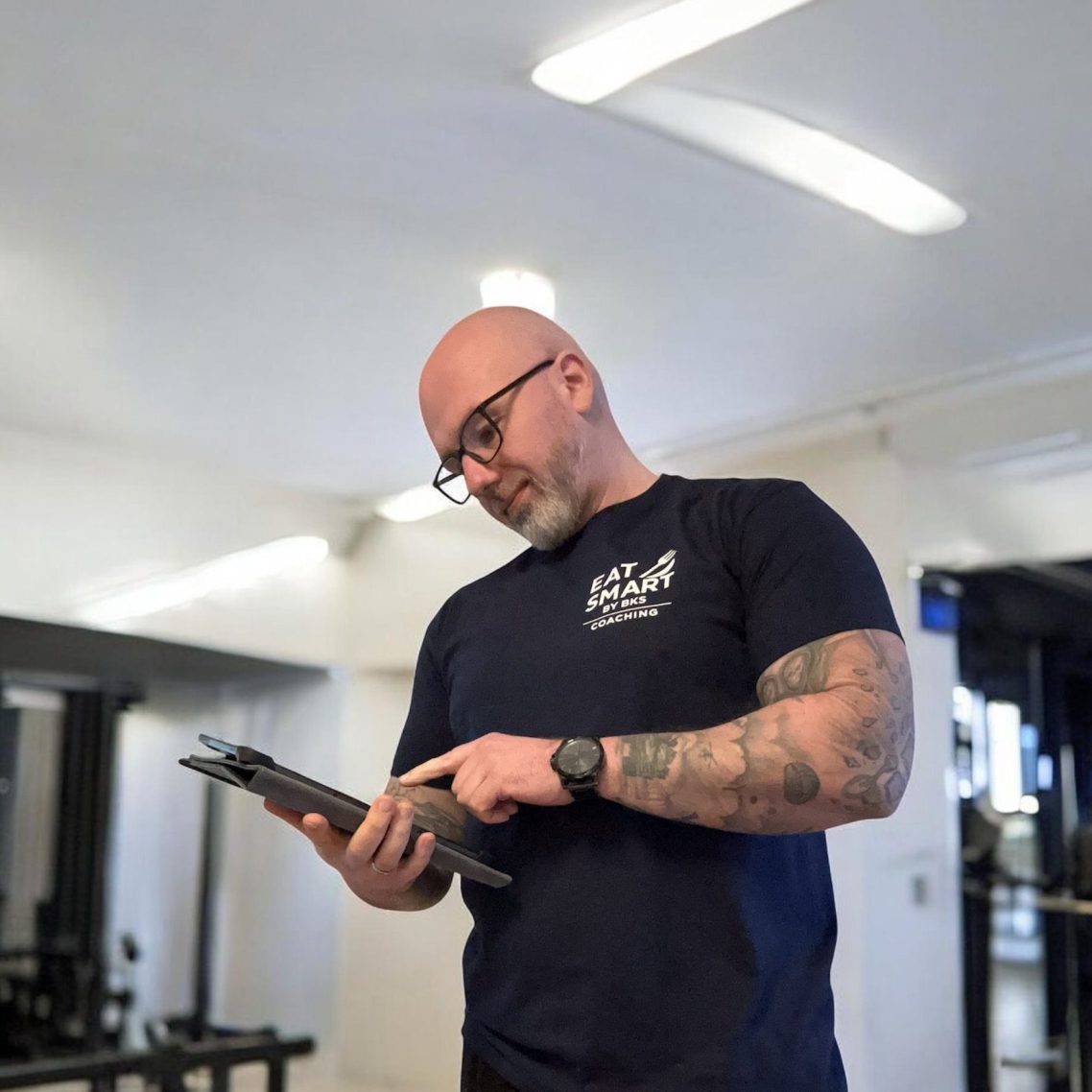 Online Coaching Lächelnder Mann in grauer Kapuzenjacke steht in einem modernen Fitnessstudio.